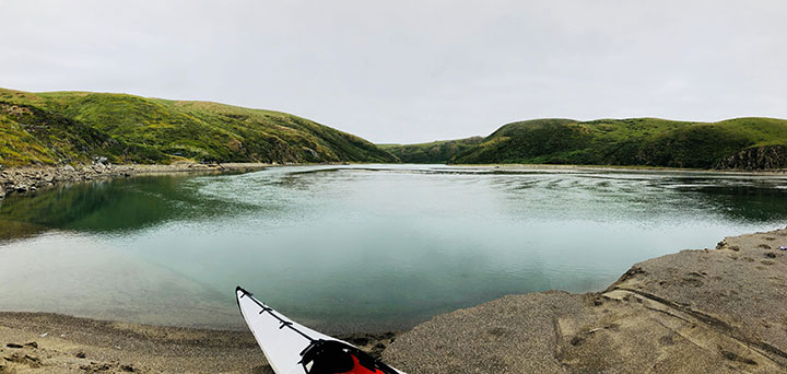 a white kayak sits on coarse sand in the foreground, green calm waters extend out to rolling green hills meeting an overcast gray sky